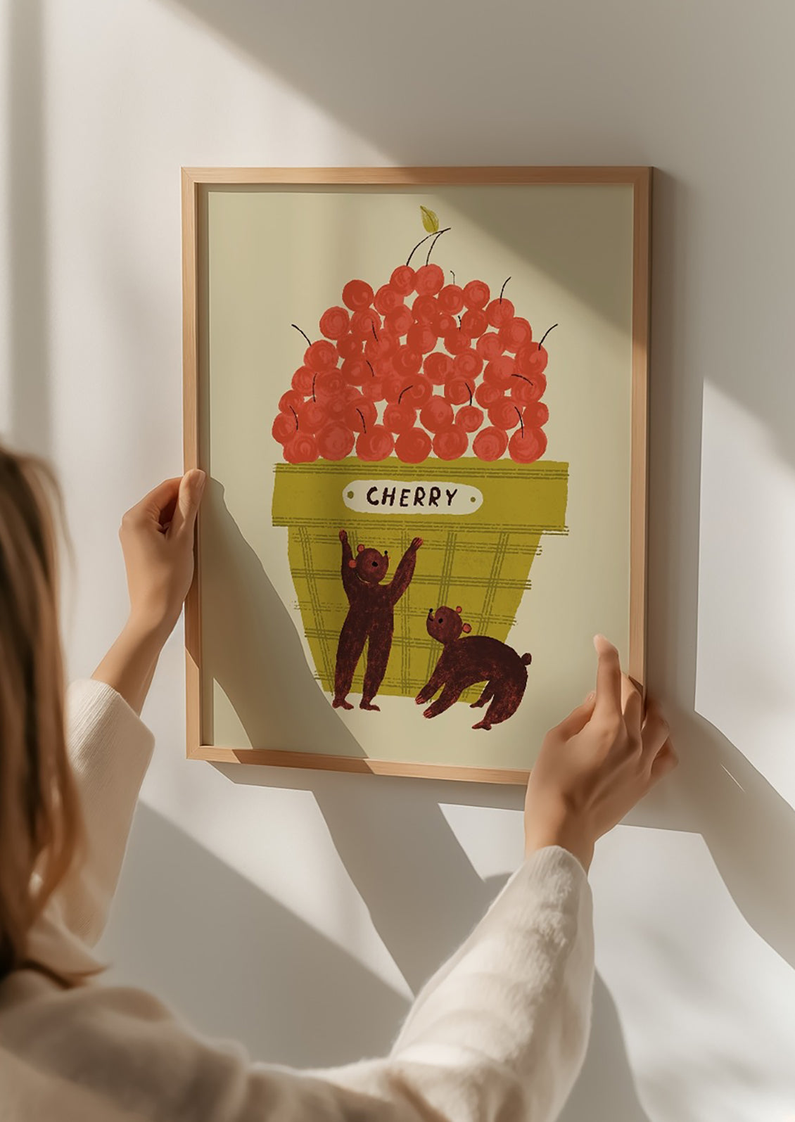 Person holding a framed illustration of cherries and bears on a white wall