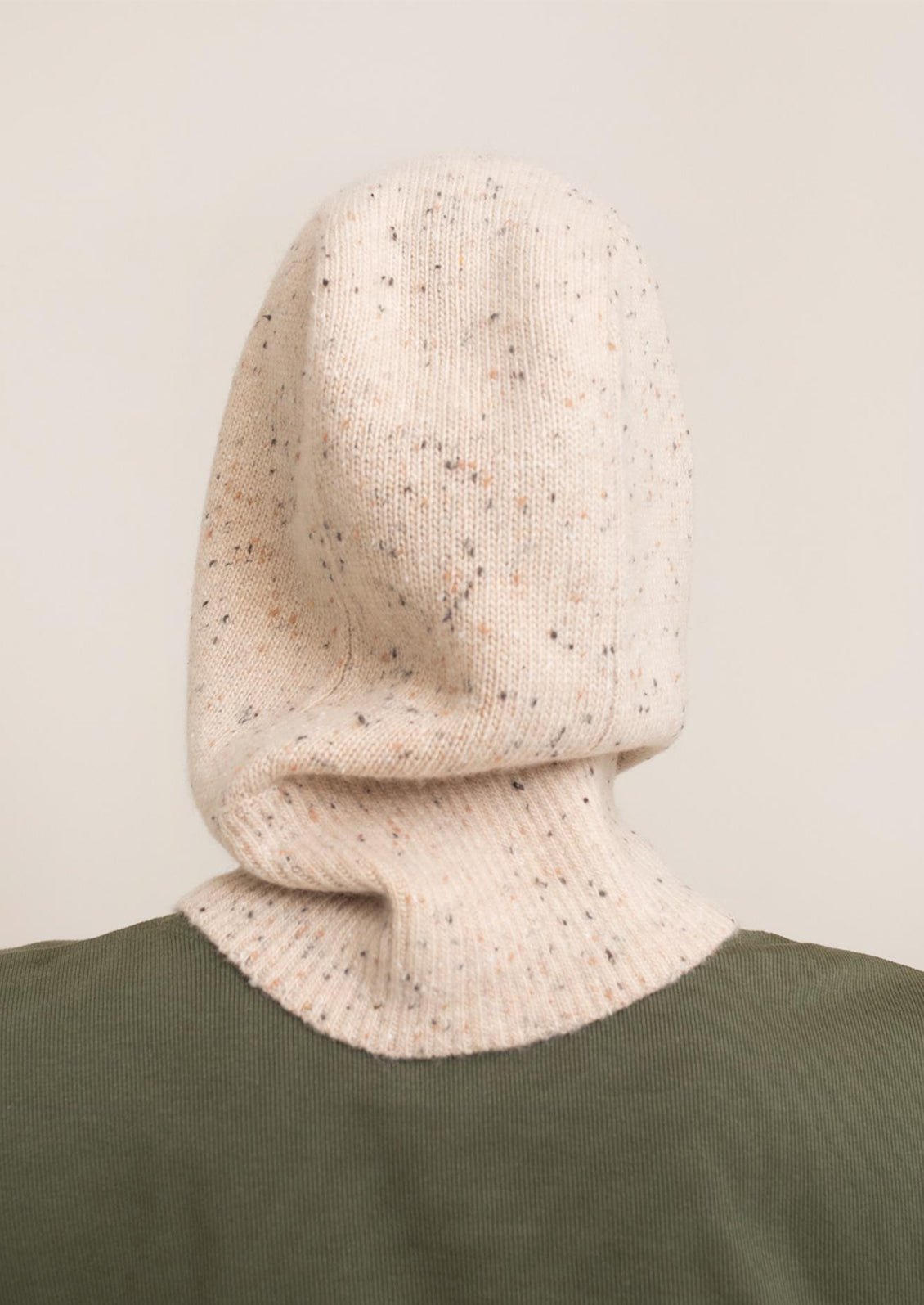 Person wearing a beige knitted hood over a green garment on a neutral background