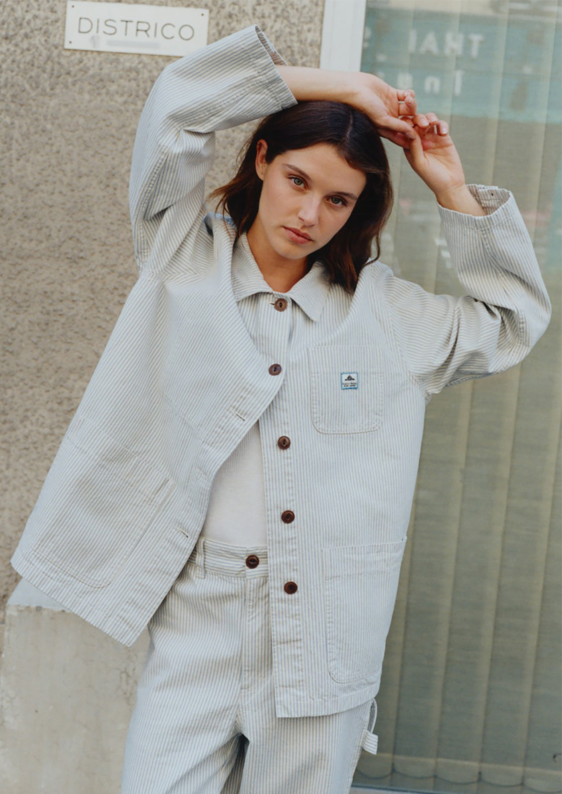 Woman wearing a light-colored jacket and pants against a textured wall.