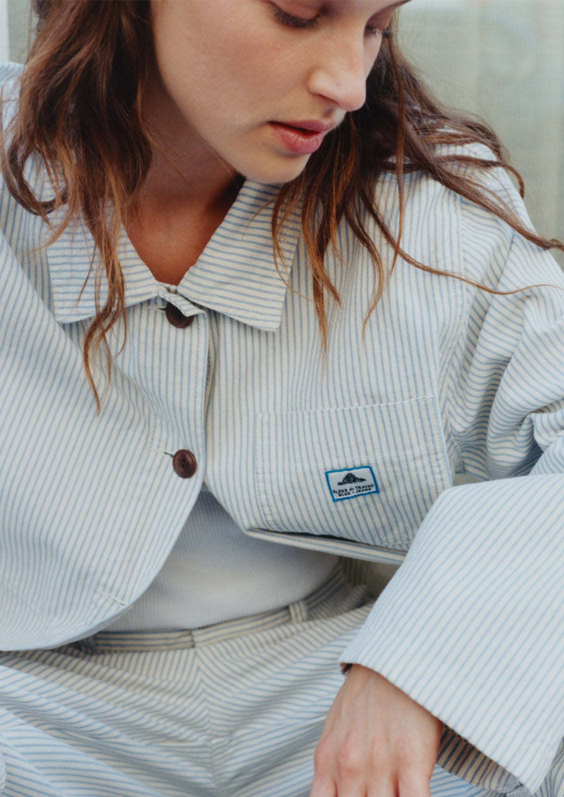 Person wearing a light gray striped jacket with a brand logo, sitting on a light-colored surface.
