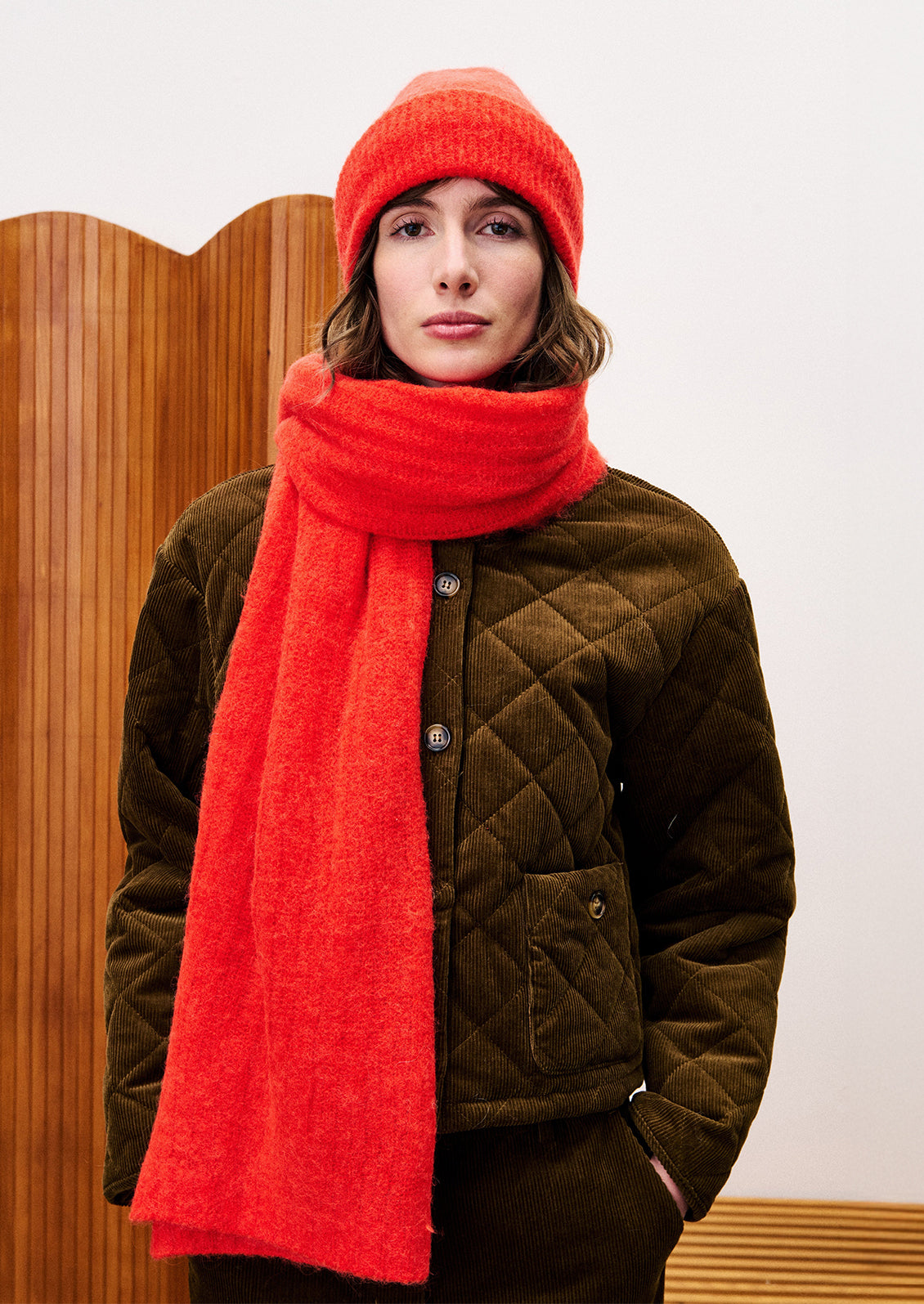 Person wearing a red scarf and beanie with a brown quilted jacket against a wooden panel background