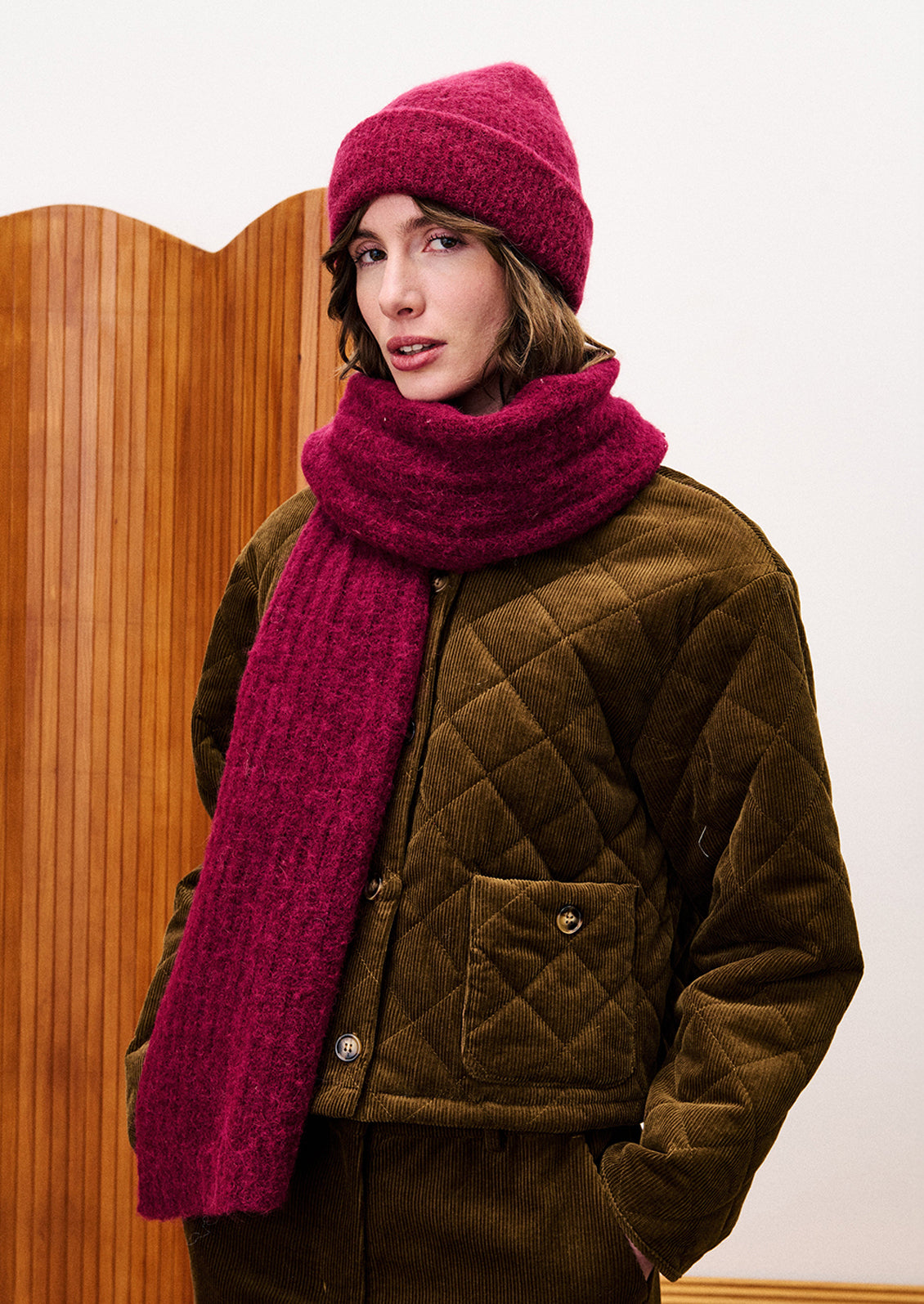 Person wearing a brown quilted jacket, pink beanie, and pink scarf against a wooden panel background