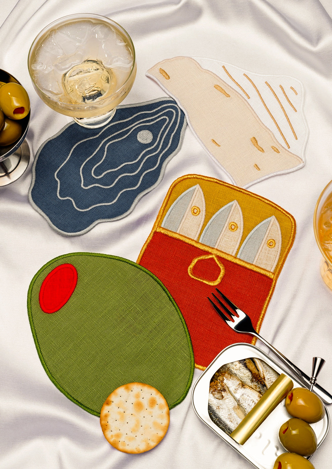 Table setting with decorative cocktail napkins shaped like an olive, an oyster, a tin of sardines and a piece of cheese.