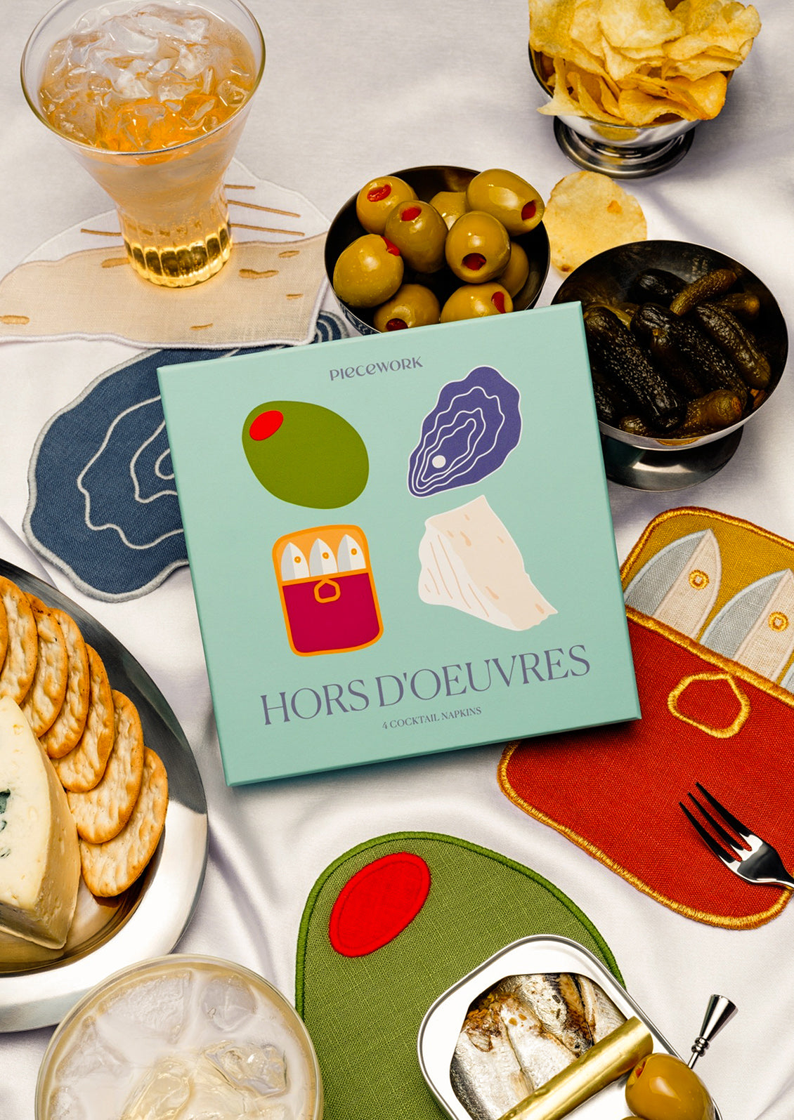 Table setting with decorative cocktail napkins shaped like an olive, an oyster, a tin of sardines and a piece of cheese.
