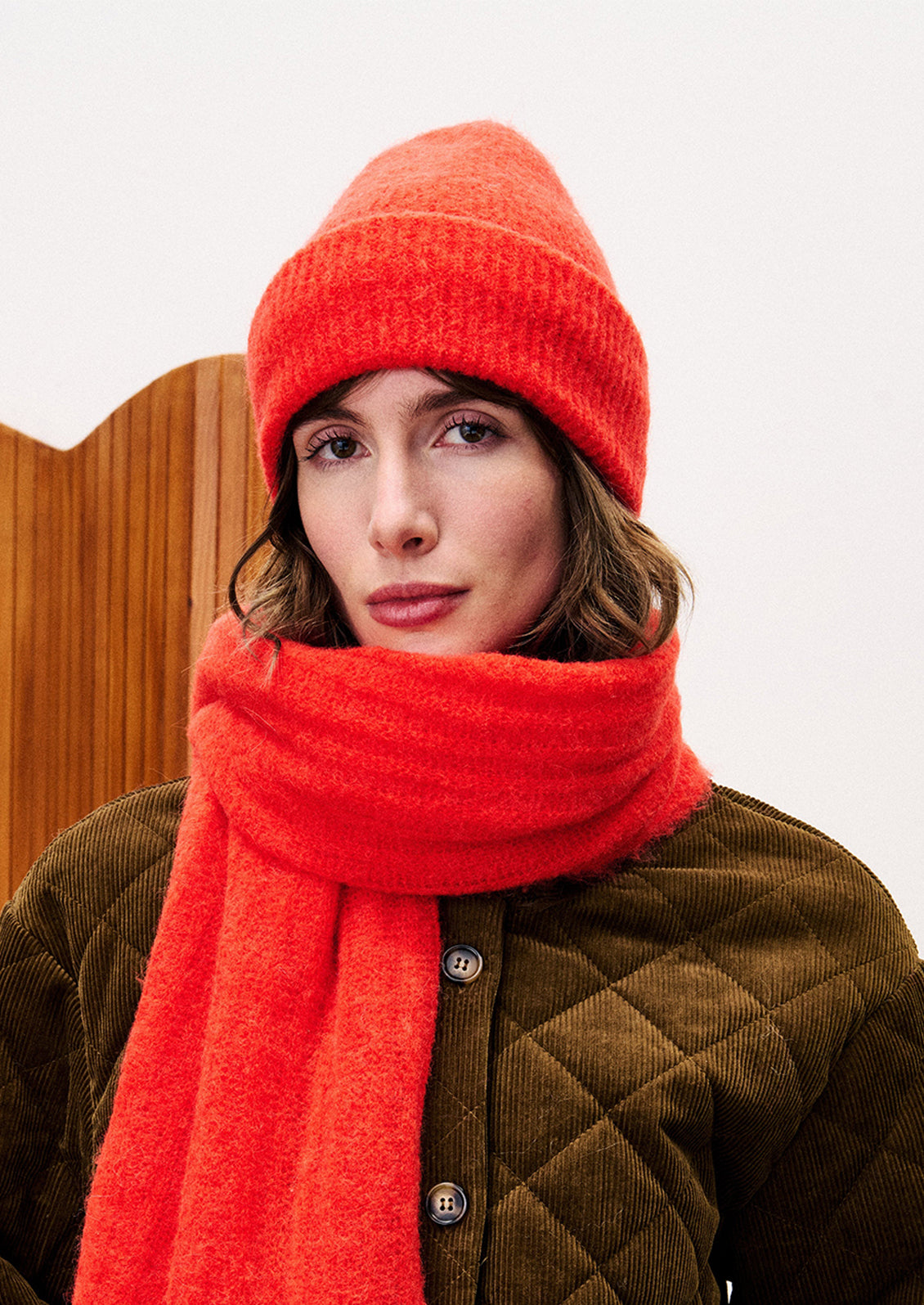 Person wearing a red knit hat and scarf with a brown quilted jacket against a white background