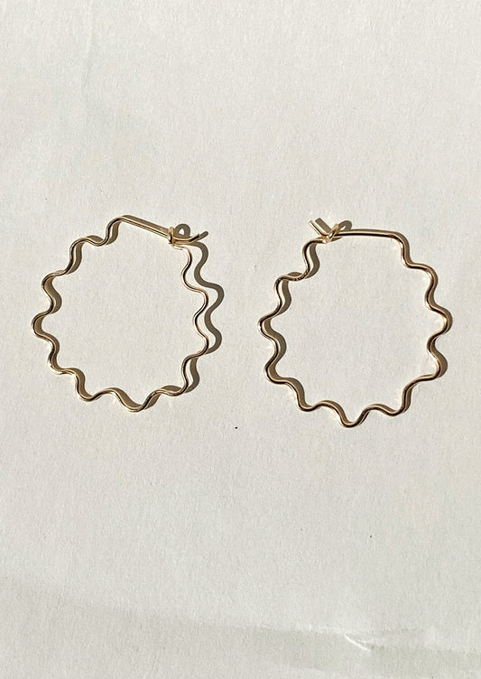 Gold hoop earrings with a wavy squiggle shape on a light beige background.