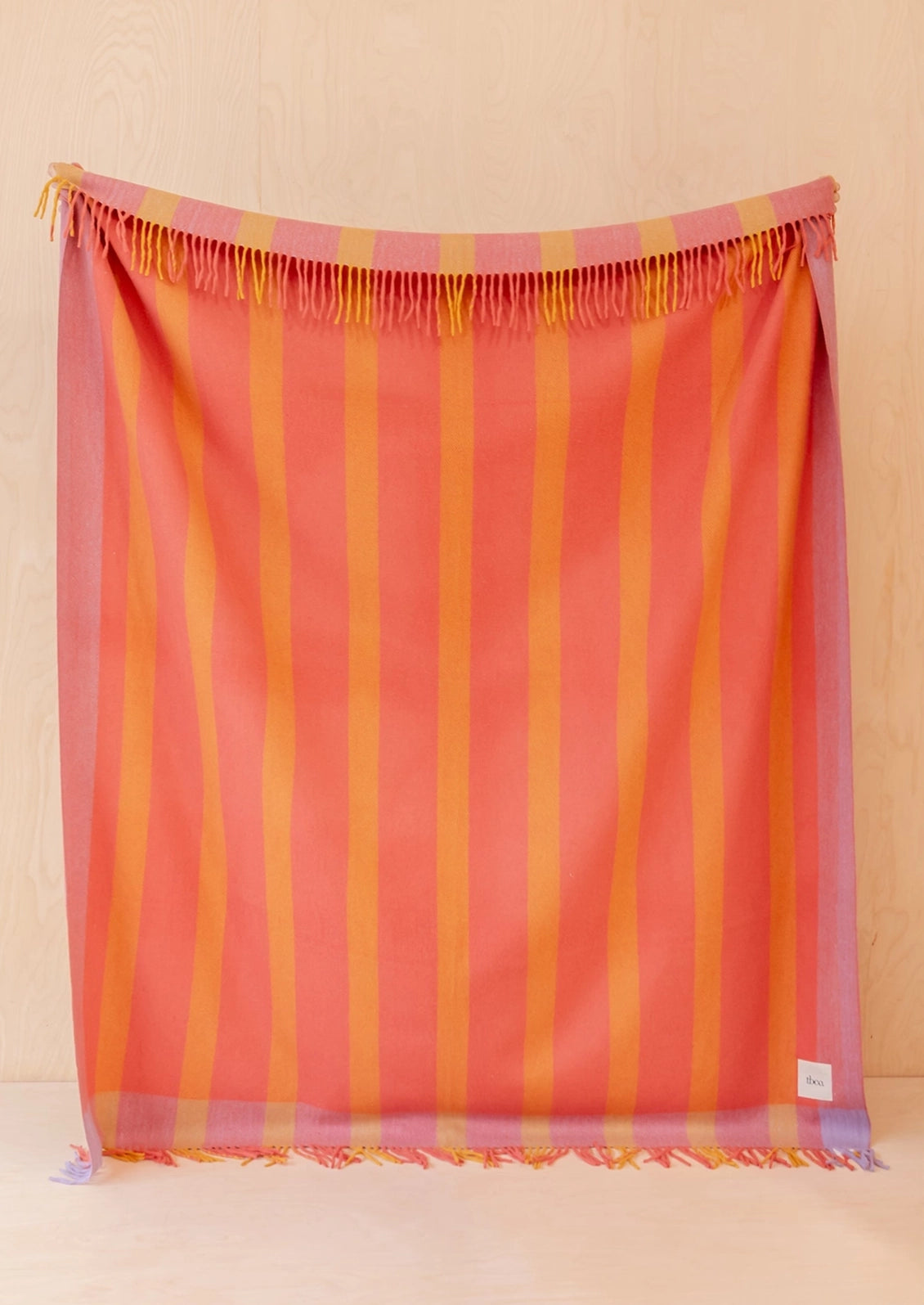 A throw blanket in shades of coral pink, orange, and yellow, featuring fringe trim along the top and bottom edges.