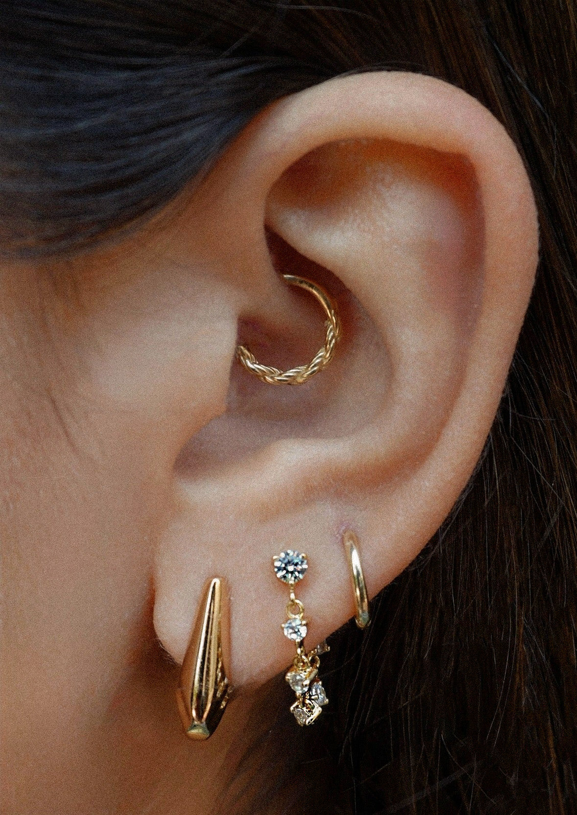 A woman wearing various gold earrings including two minimal hoop earrings and a gold chain stud earring adorned with cubic zirconia gems.