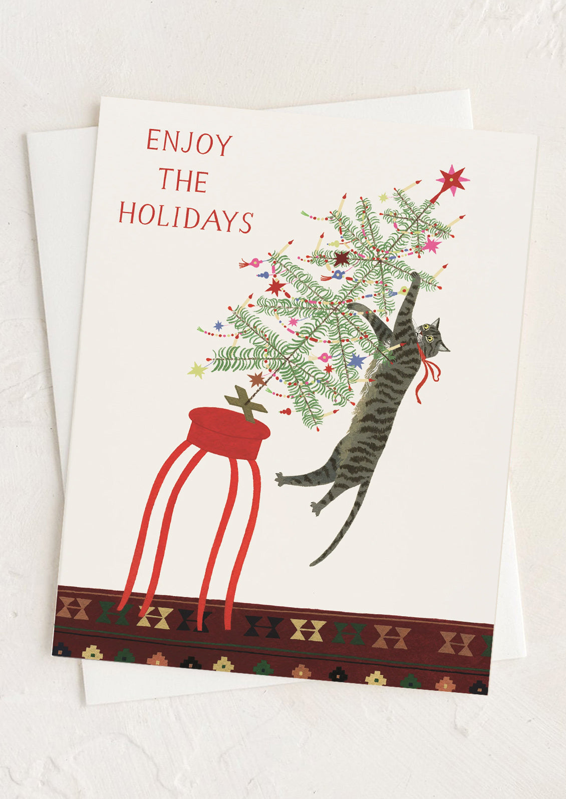 A greeting card with illustration of a cat swinging off of a small Christmas tree, text reads "Enjoy the holidays".