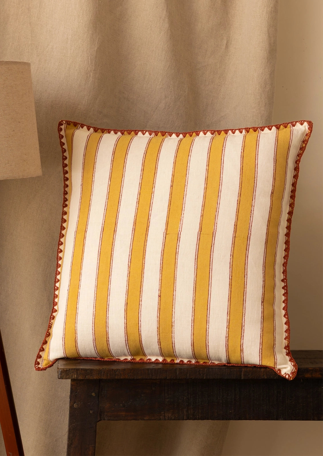 A square decorative throw pillow with vertical yellow and off-white stripes and a red zigzag border, resting on a dark wooden table against a beige curtain background.