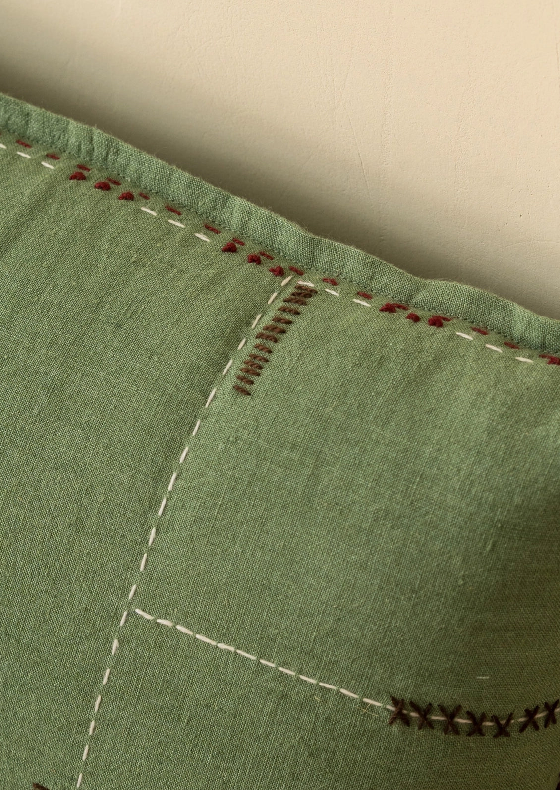 Close-up of a sage green fabric cushion or pillow with decorative hand-stitched seams in white, red, and dark brown.