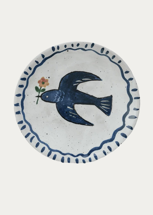 A small ceramic plate with hand painted imagery of blue bird holding flower.