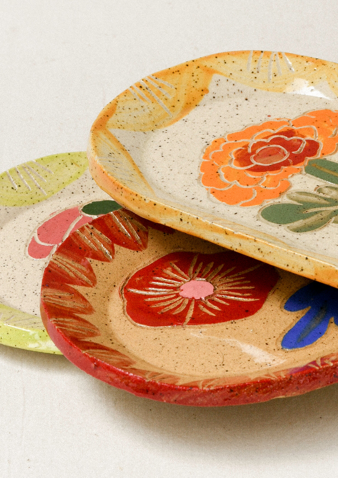 Three ceramic oval catchall trays stacked on top of on another, each with an etched flower in the center and colorful border.