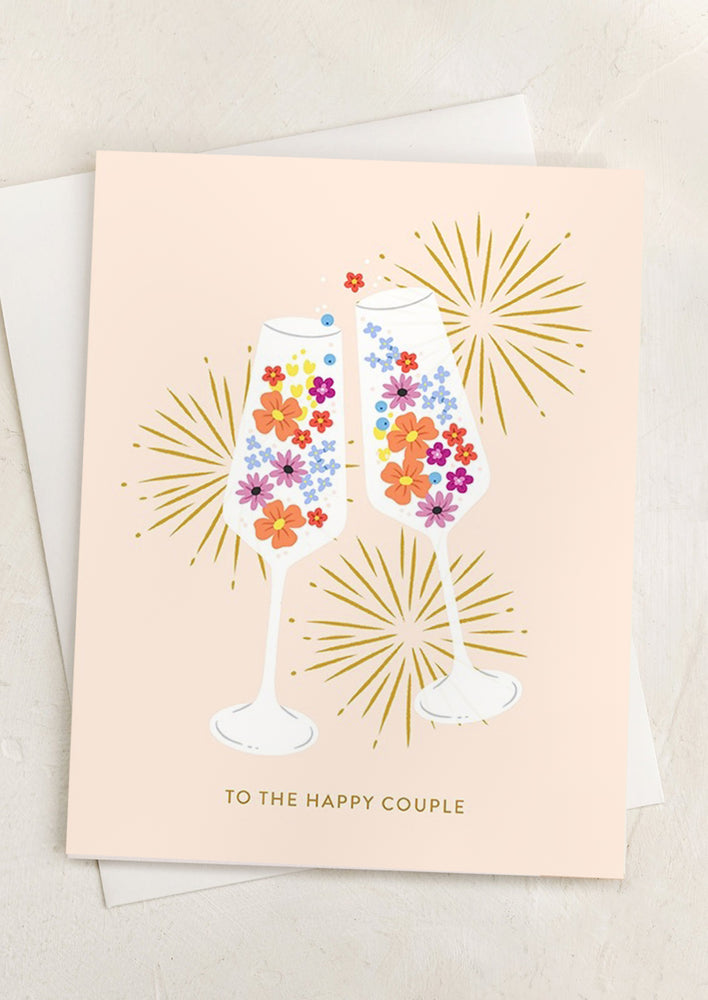 A peach colored card with illustration of clinking champagne glasses full of flowers, text reads "To the happy couple".