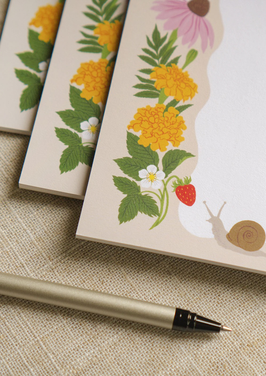 Closeup of three notepads stacked in a staggered fashion with a ripple white section and an illustration of a happy snail and ladybug enjoying yarrow, marigold, coneflower, and fresh strawberries.