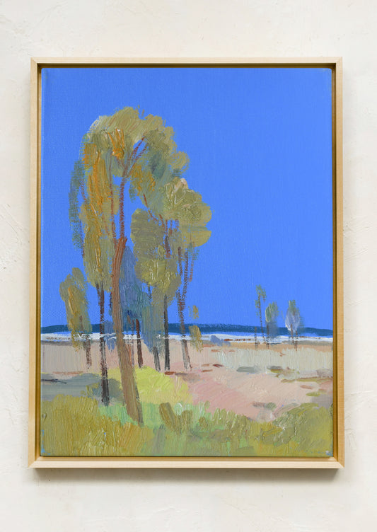 A framed landscape painting featuring tall green trees under a bright blue sky, hung on a textured off-white wall.