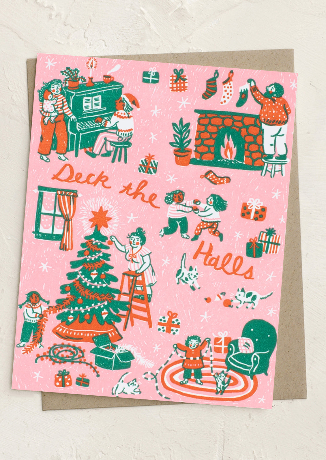 A pink Christmas greeting card with a whimsical hand-drawn illustration of holiday scenes, including a decorated tree, fireplace, stockings, gifts, and people, with the words 'Deck the Halls' in orange script. The card is shown on top of a brown envelope against a light textured background.
