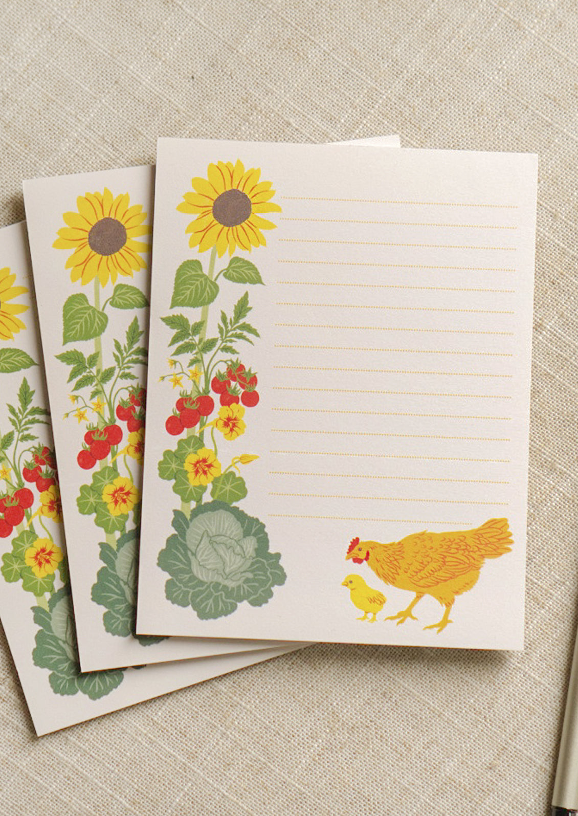 Three notepads stacked in a staggered fashion featuring ripple dotted lines and an illustration of a mother hen and baby chick exploring a vegetable garden with a large sunflower and tomatoes.