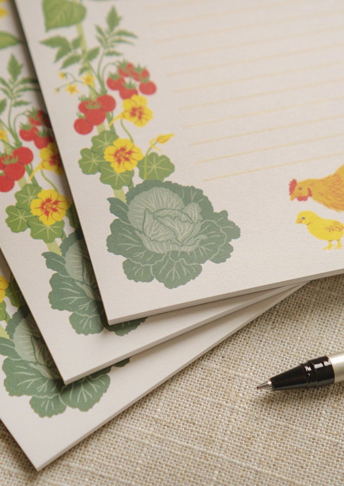 Closeup of three notepads stacked in a staggered fashion featuring ripple dotted lines and an illustration of a mother hen and baby chick exploring a vegetable garden with a large sunflower and tomatoes.