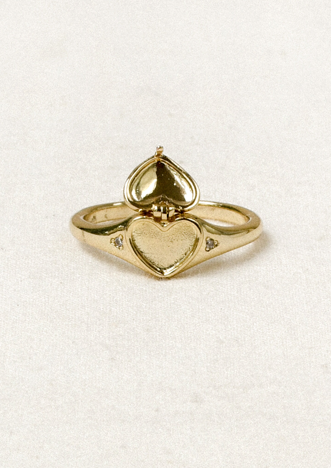 A gold ring with a tiny open locket in the shape of a heart in the center and crystal detailing on either side.