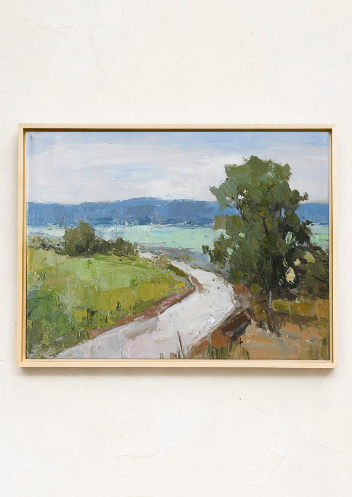 A framed landscape oil painting on a textured off-white wall. The painting depicts a winding light-colored path through green fields and trees towards blue hills under a pale sky, enclosed in a light natural wood frame.