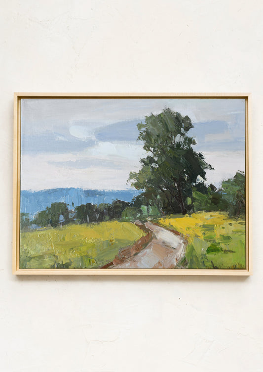 A framed landscape painting on a textured off-white wall, depicting a winding dirt path through a green field under a cloudy blue sky.
