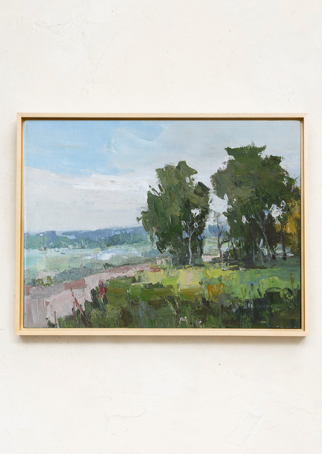 A framed landscape oil painting featuring green trees and a field under a light blue sky, mounted on a textured off-white wall.