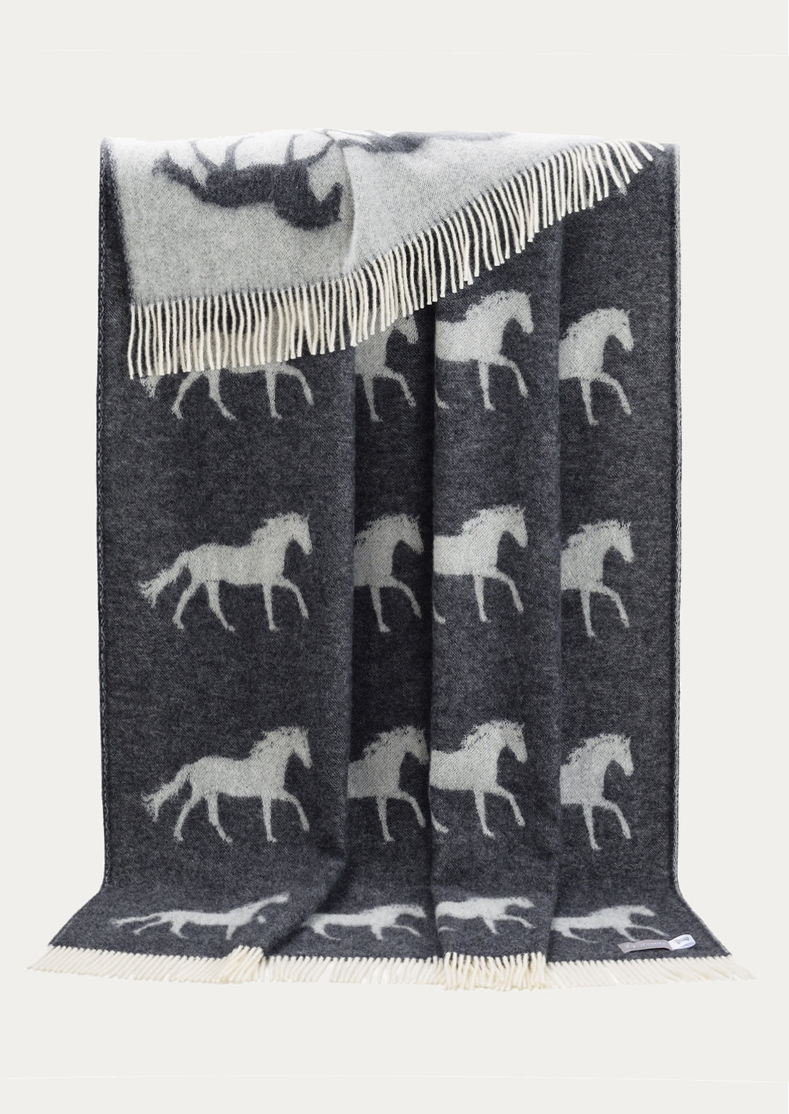 A dark gray woven throw blanket with white horse silhouettes and cream fringe, draped against a light off-white background.