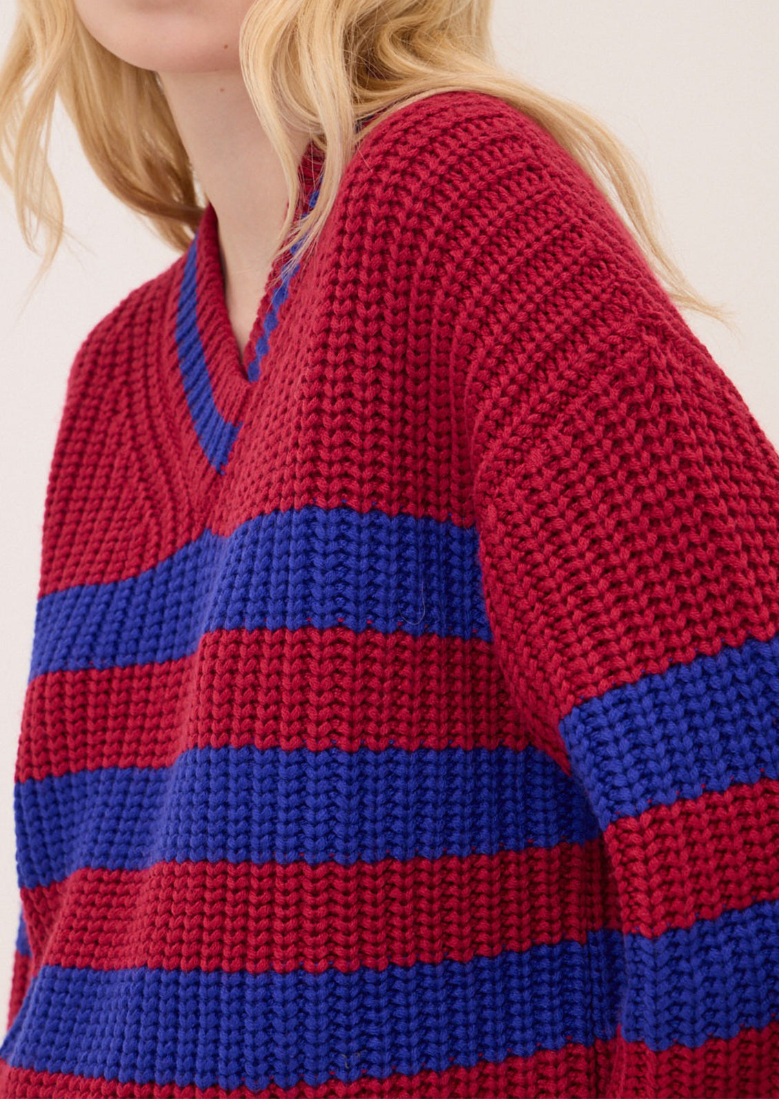 Close-up of a person wearing a chunky knit V-neck sweater with horizontal red and blue stripes, against a plain light background.