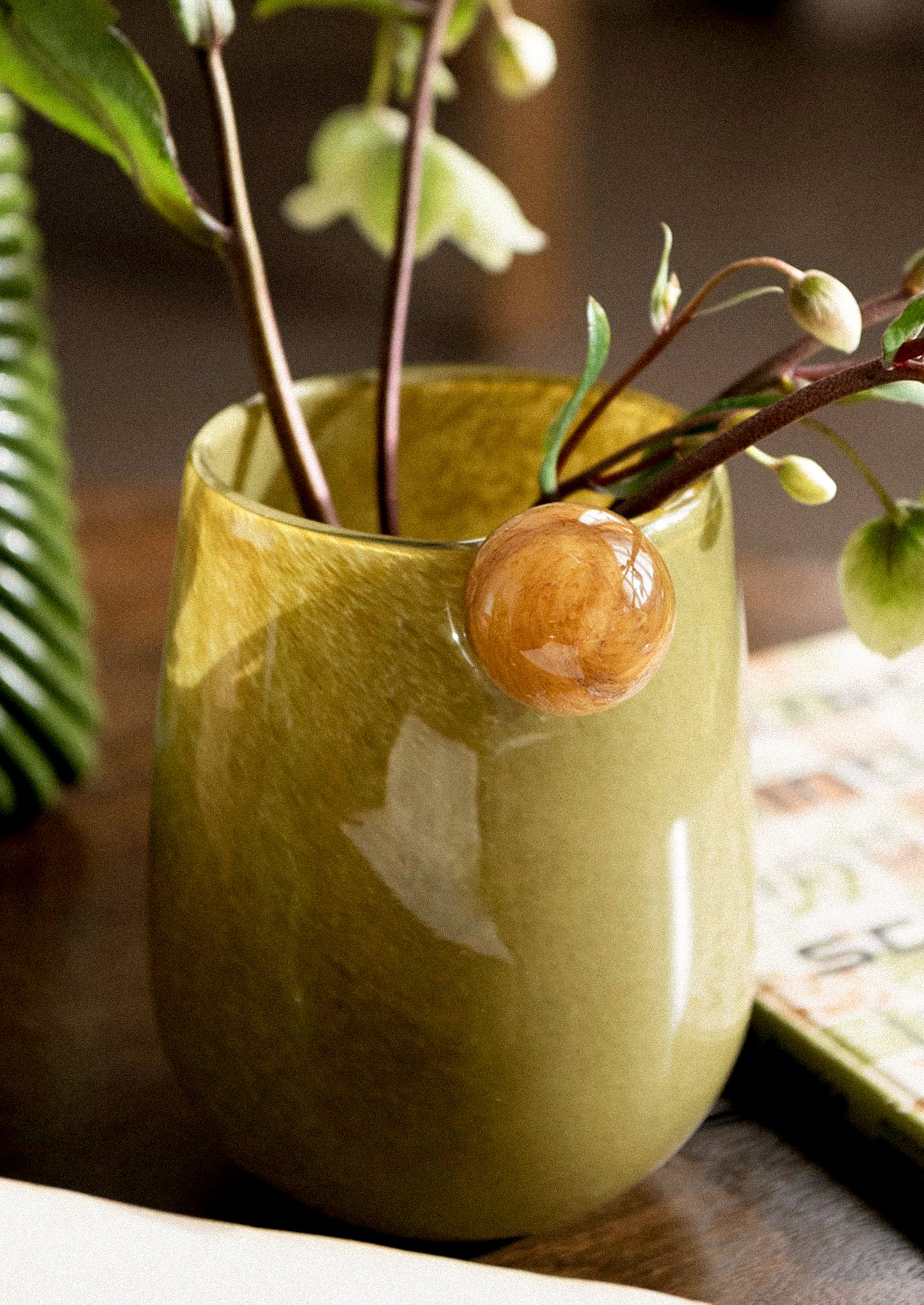 A glossy olive green glass vase with a small contrast colored ball handle on the side.