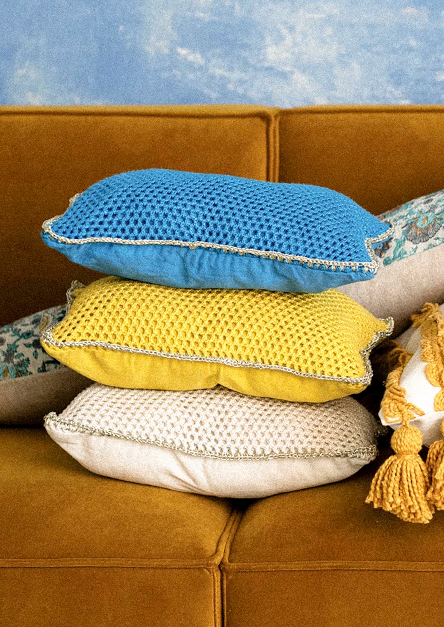 Stack of textured pillows in blue, yellow, and white on a brown couch with a blue textured wall in the background.