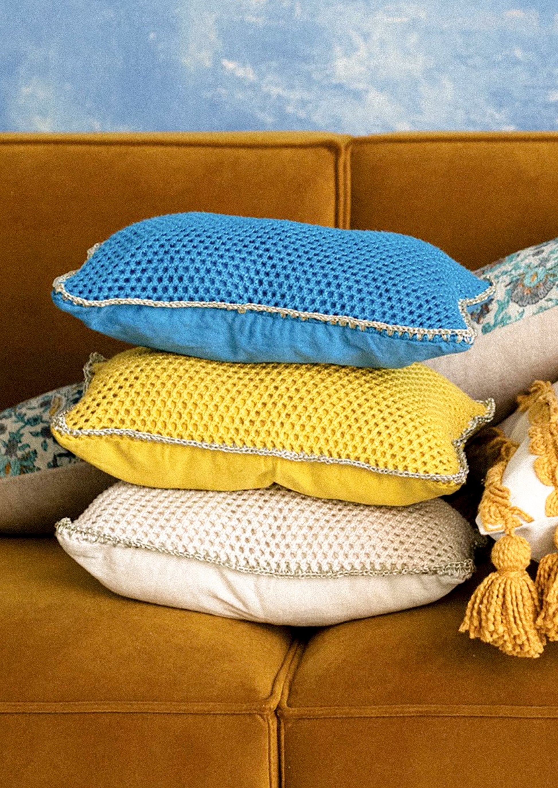 Stack of textured pillows in blue, yellow, and white on a brown couch with a blue textured wall in the background.