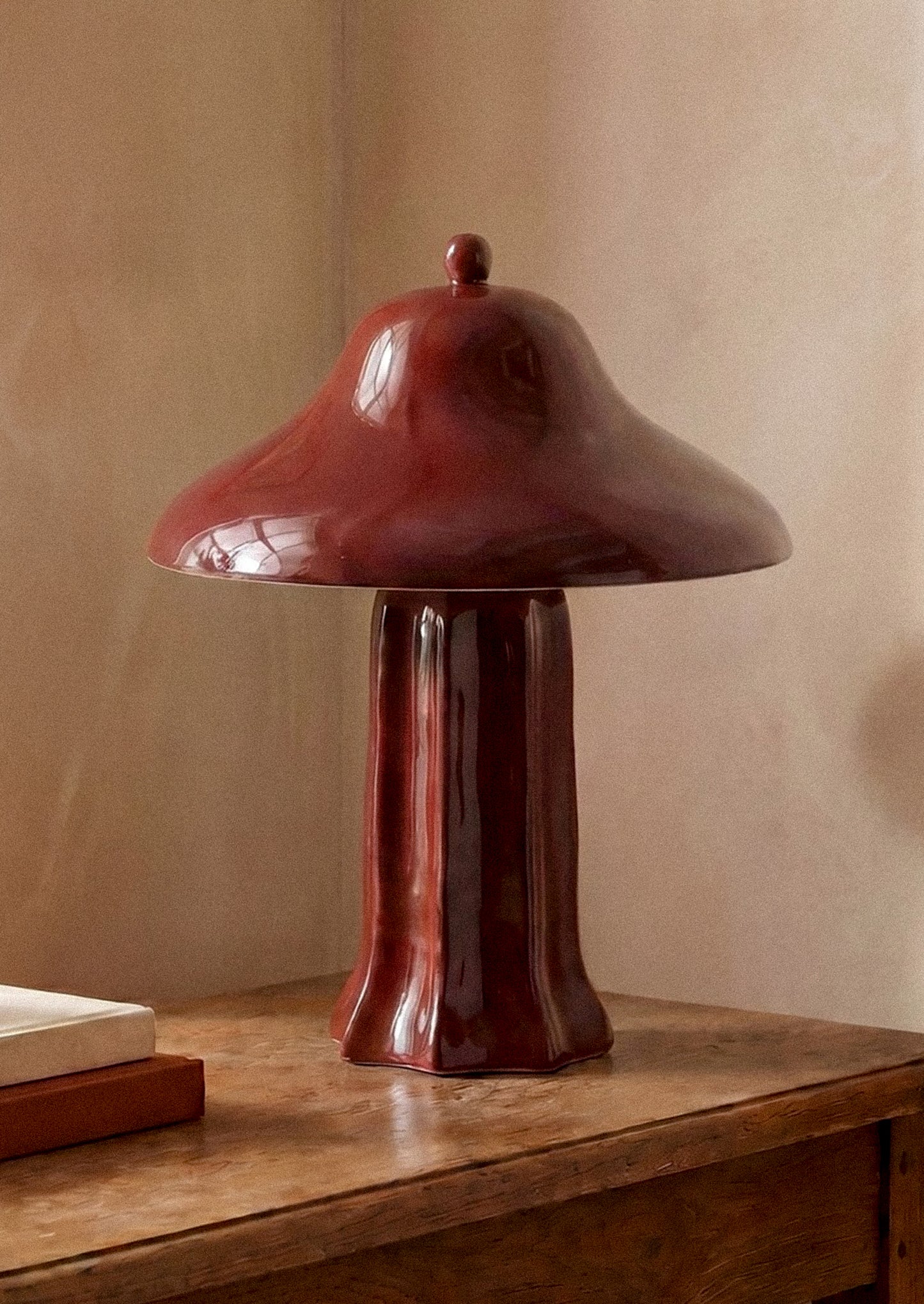 A glossy dark red ceramic table lamp with a mushroom-shaped shade and pedestal base, displayed on a wooden table against a warm beige wall.