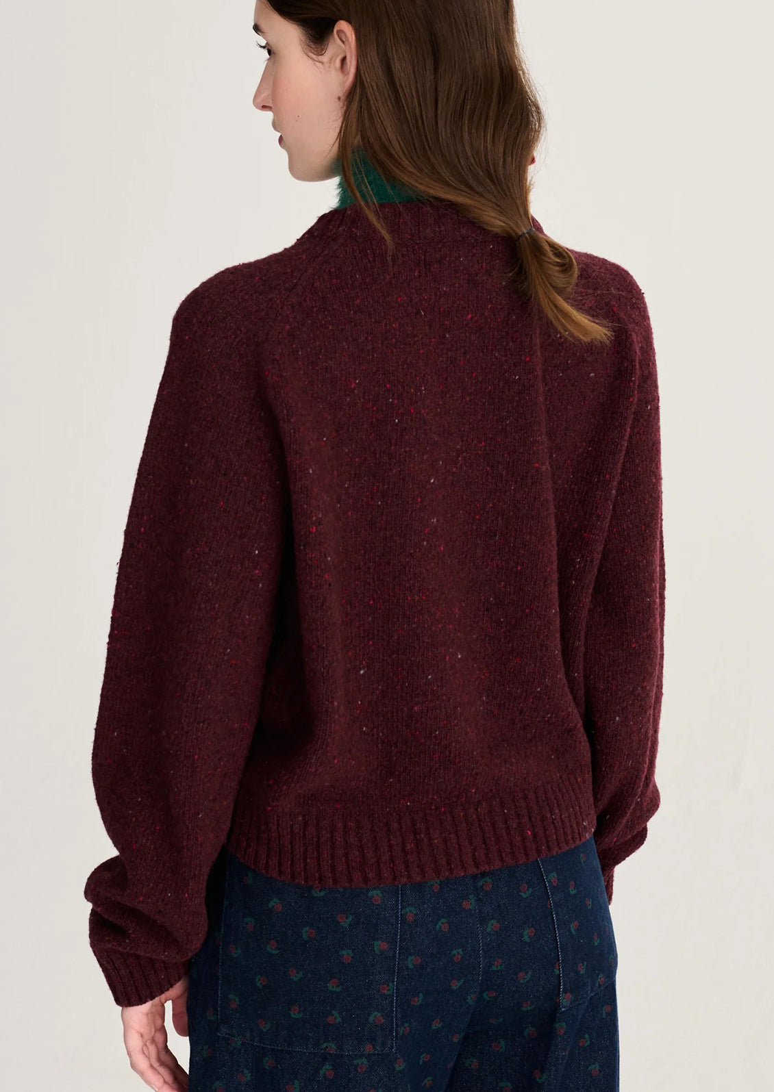A woman shown from the back wearing a dark burgundy knit sweater with subtle flecks and navy patterned trousers, against a plain off-white studio background.