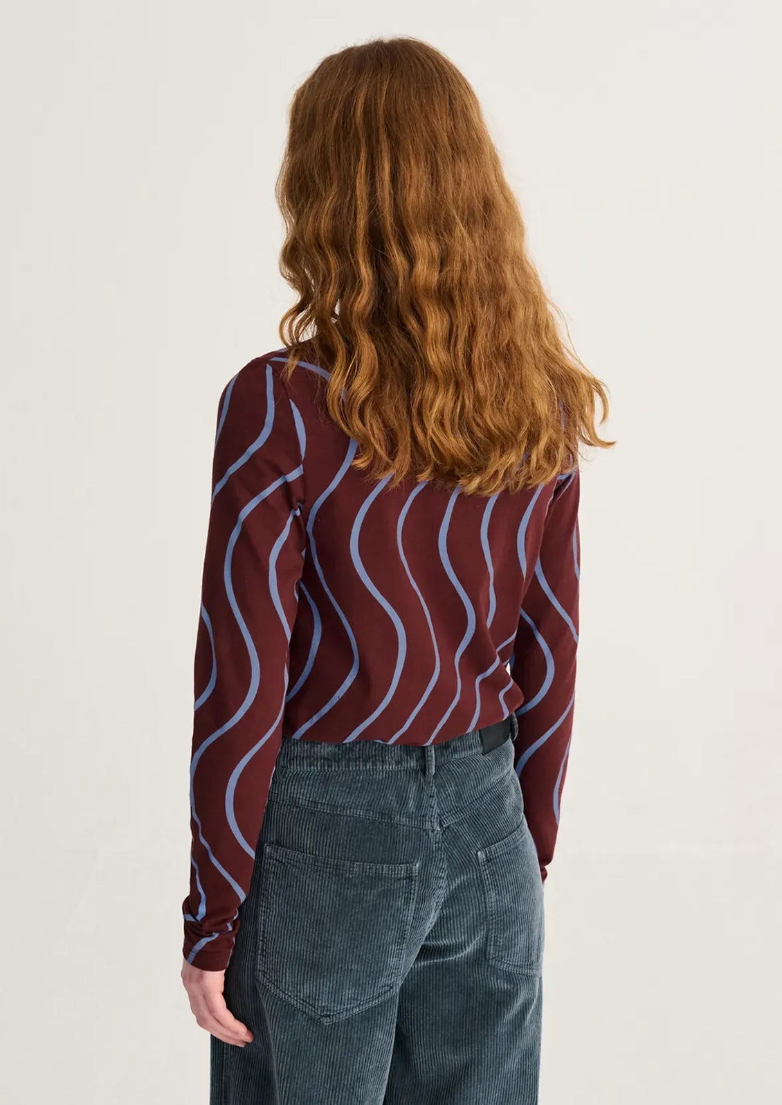 A woman with long wavy auburn hair is shown from behind wearing a long-sleeve top with a burgundy base and light blue wavy stripes, paired with dark blue jeans, against a plain off-white background.