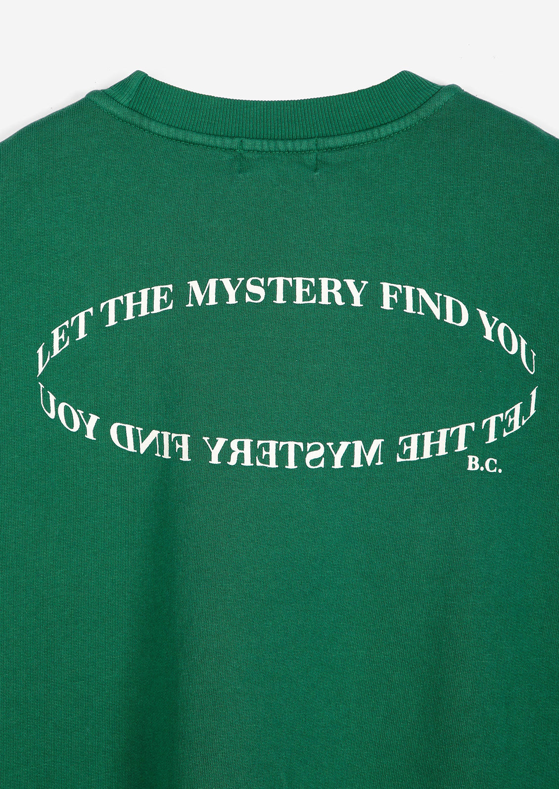 A close-up of a green crewneck sweatshirt showing the back, which features a white graphic text design that reads 'LET THE MYSTERY FIND YOU' in an oval shape, with 'B.C.