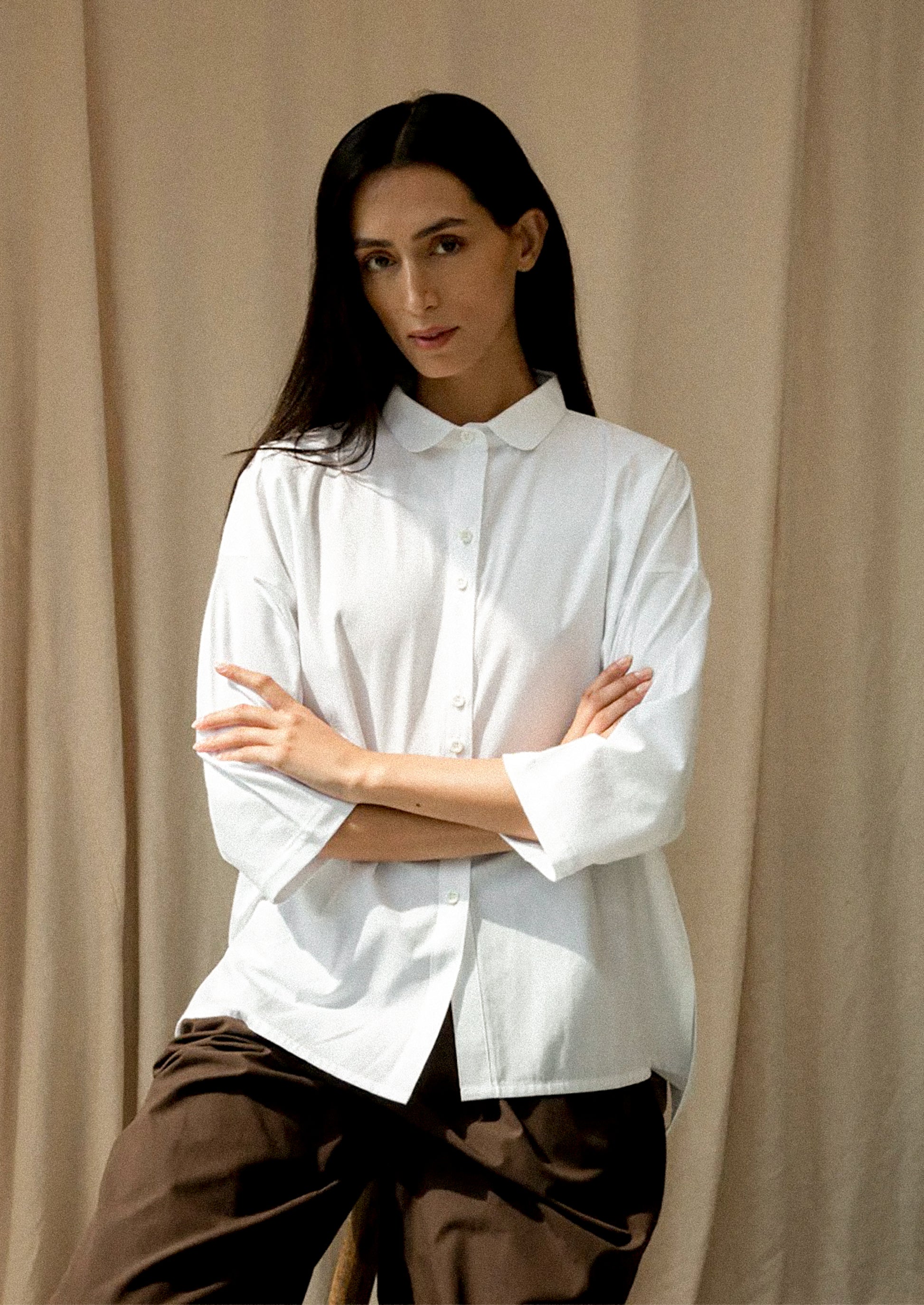 Person wearing a white shirt with arms crossed against a beige curtain background