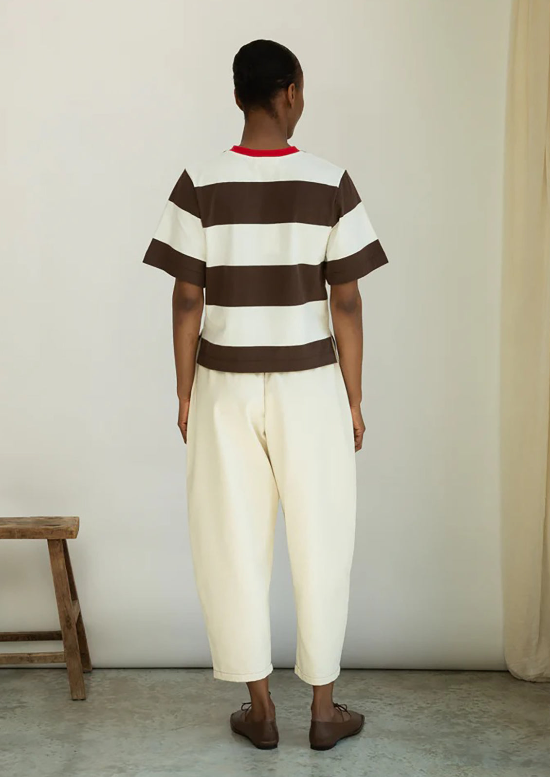 Person wearing a brown and white striped shirt and cream pants standing against a plain wall.