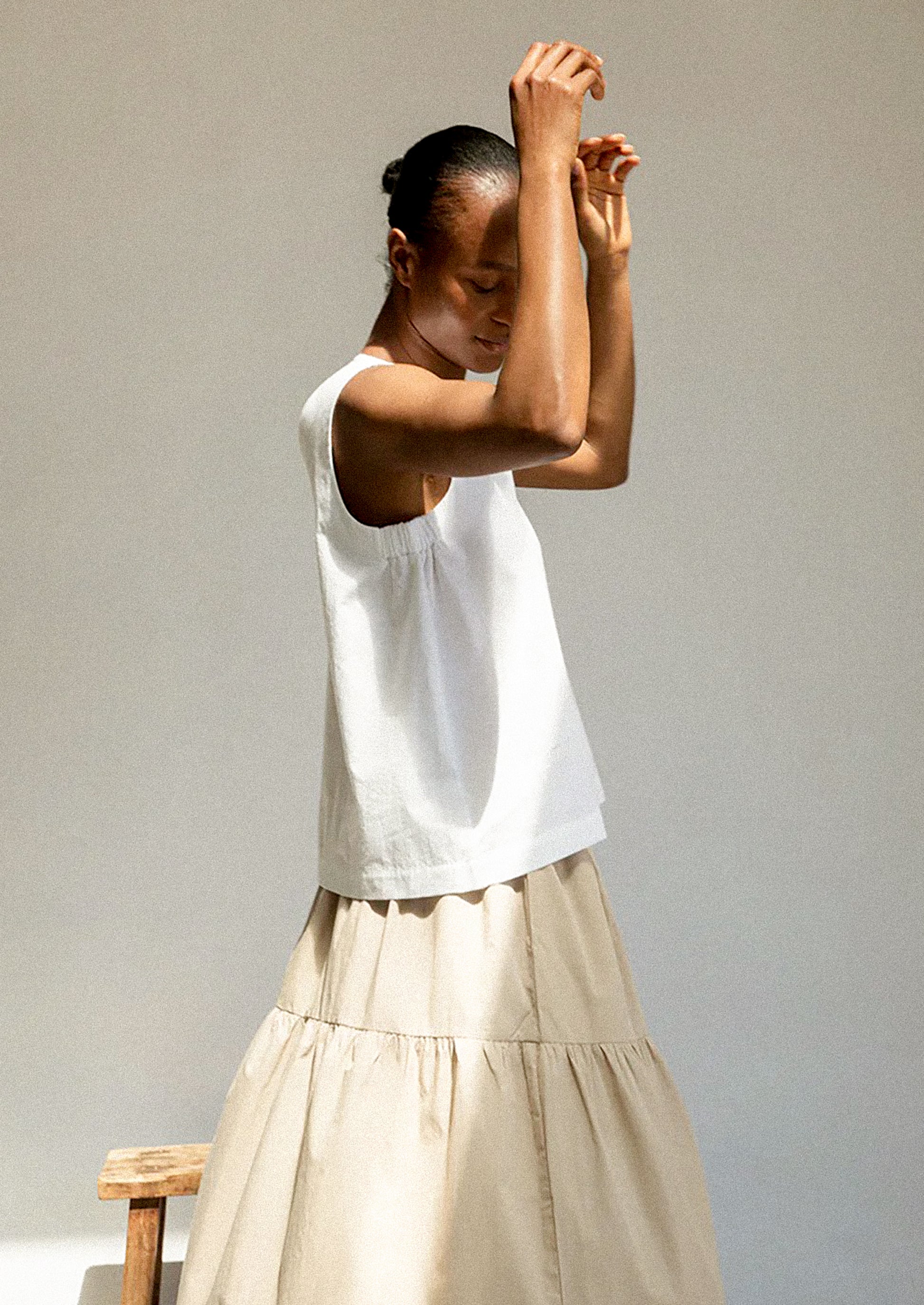 Woman wearing a white sleeveless top and beige skirt against a plain background