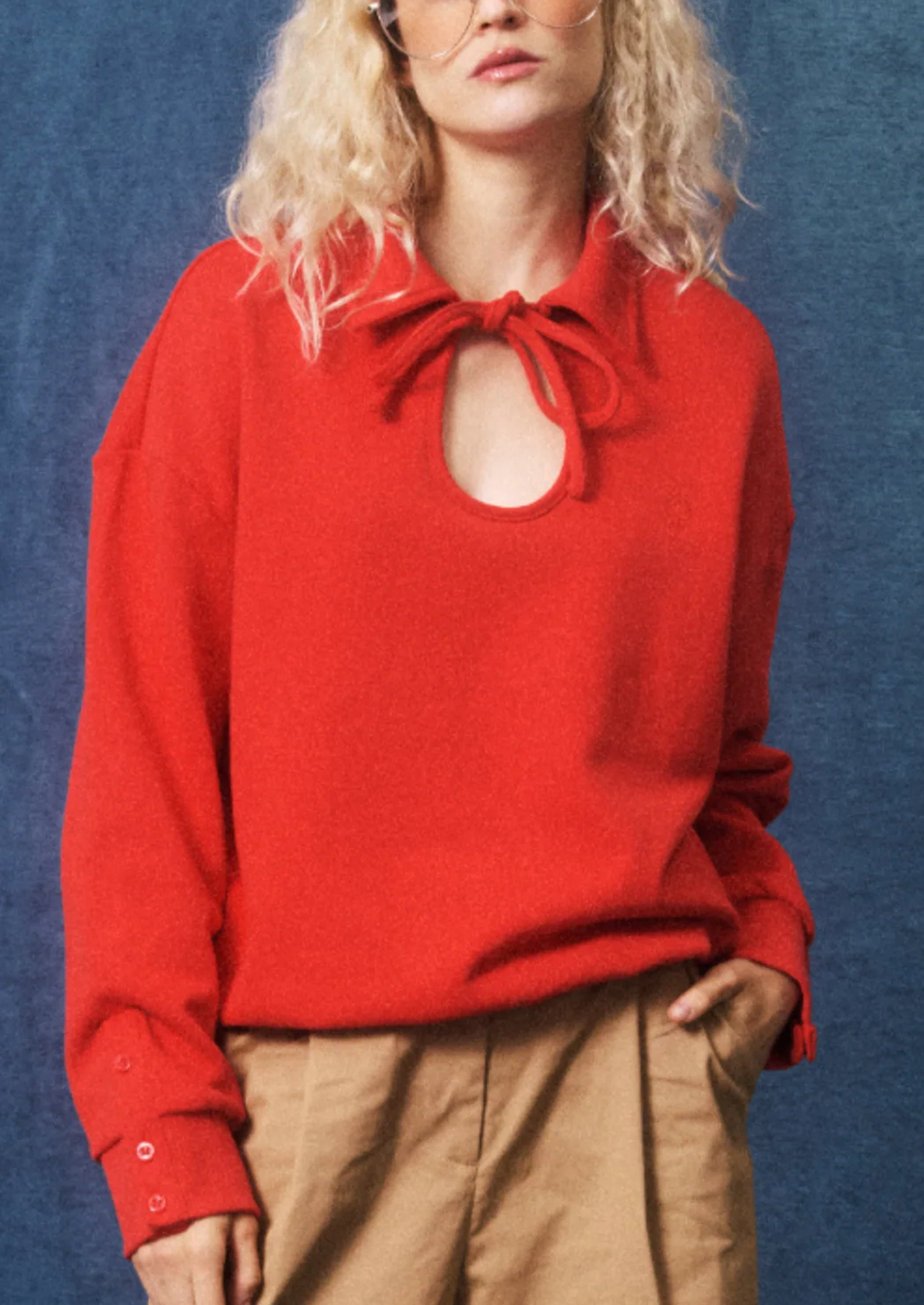 A woman wearing a bright red knit sweater with a keyhole cutout and tie-neck detail, paired with tan trousers, set against a textured blue backdrop.