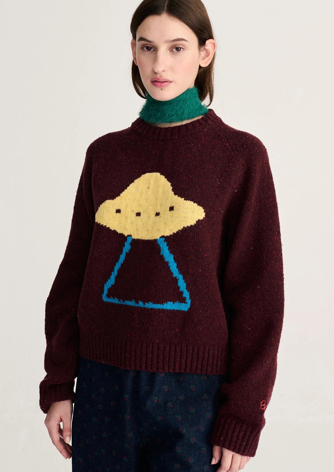 A model wearing a dark burgundy knit sweater featuring a large yellow and blue UFO graphic on the front, styled with a green turtleneck and dark patterned trousers.