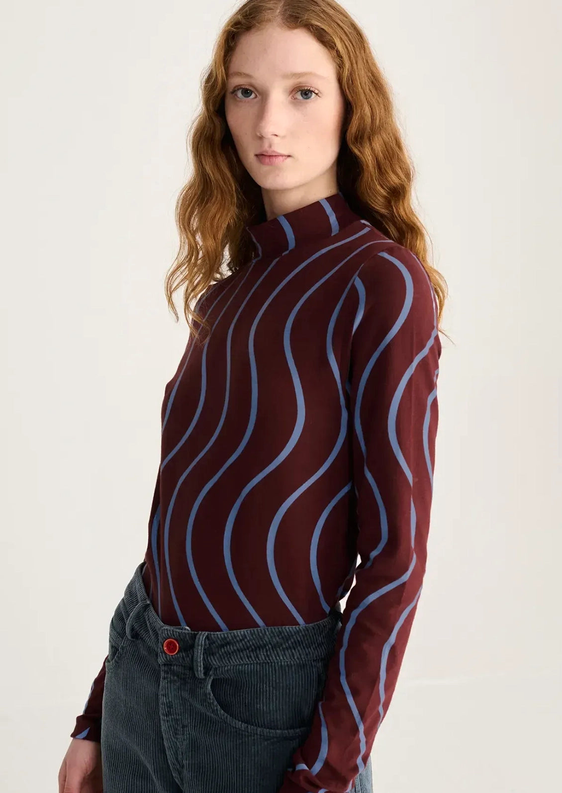 A woman with red hair wearing a long-sleeve mock neck top with a burgundy base and light blue wavy vertical stripes, paired with dark gray jeans, against a plain off-white background.
