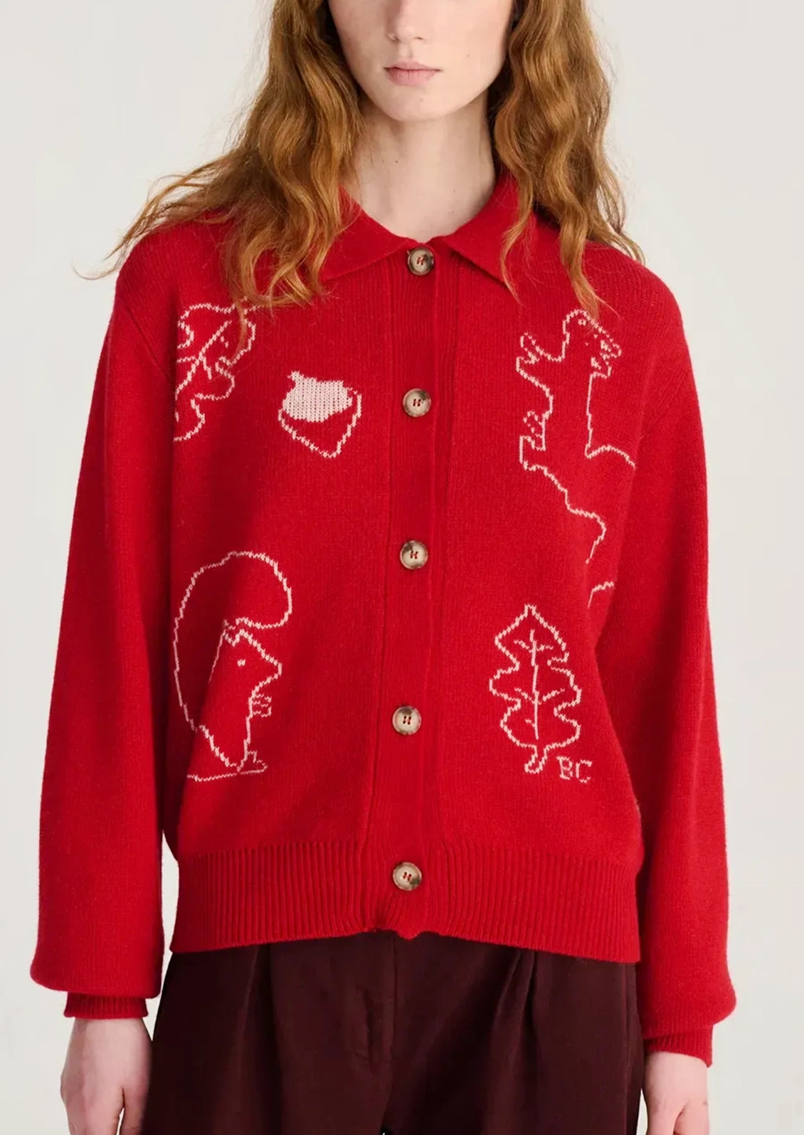 A red knit cardigan sweater with white knitted animal and leaf motifs, featuring gold buttons and a collar, worn by a model.