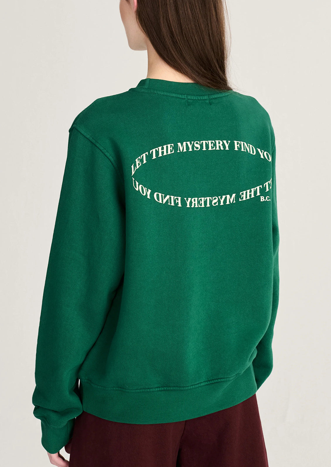 A person shown from the back wearing a dark green crewneck sweatshirt with white text on the back reading 'LET THE MYSTERY FIND YOU' and 'BY THE MYSTERY FIND YOU' in a circular design, paired with burgundy pants against a light gray background.