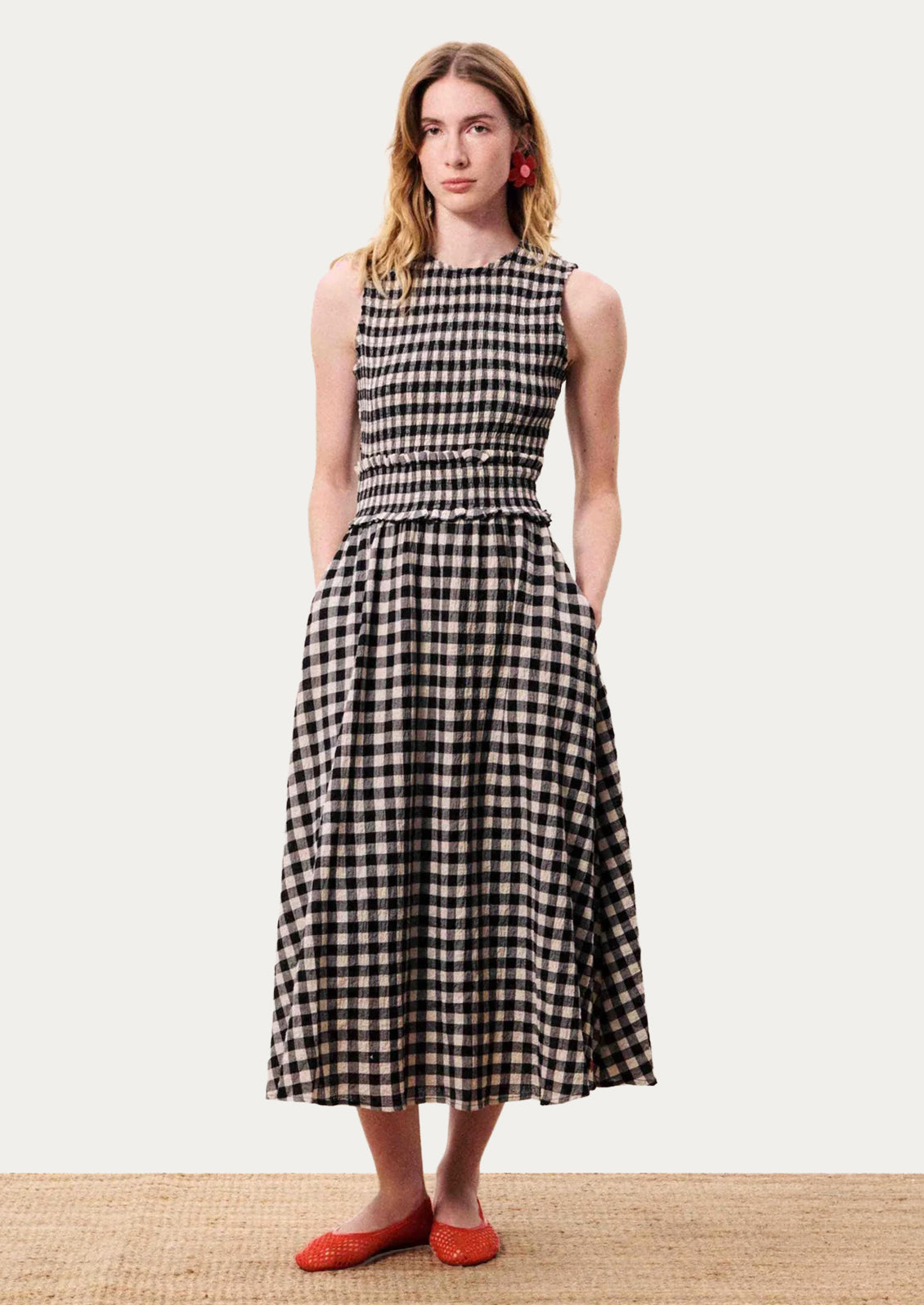 A woman modeling a sleeveless black and white gingham midi dress with a smocked waist and side pockets, paired with red shoes.