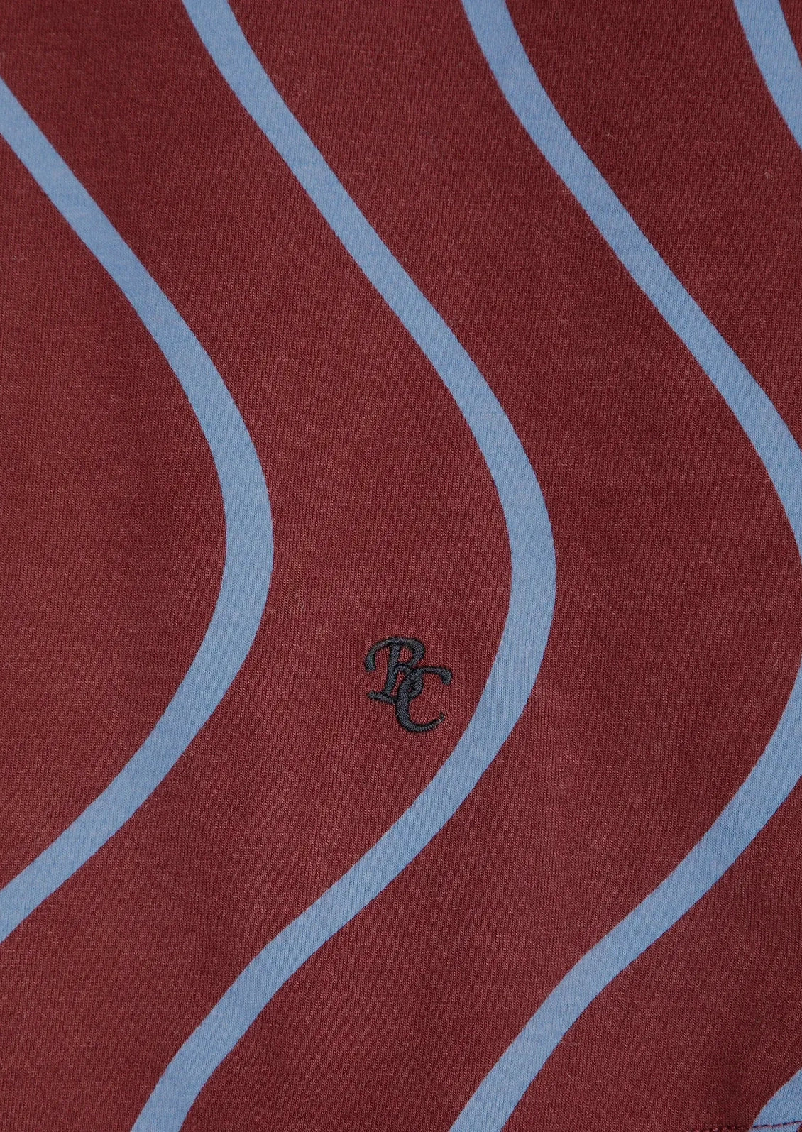 A close-up of a red fabric with a repeating light blue wavy stripe pattern and a small dark embroidered monogram logo near the center.