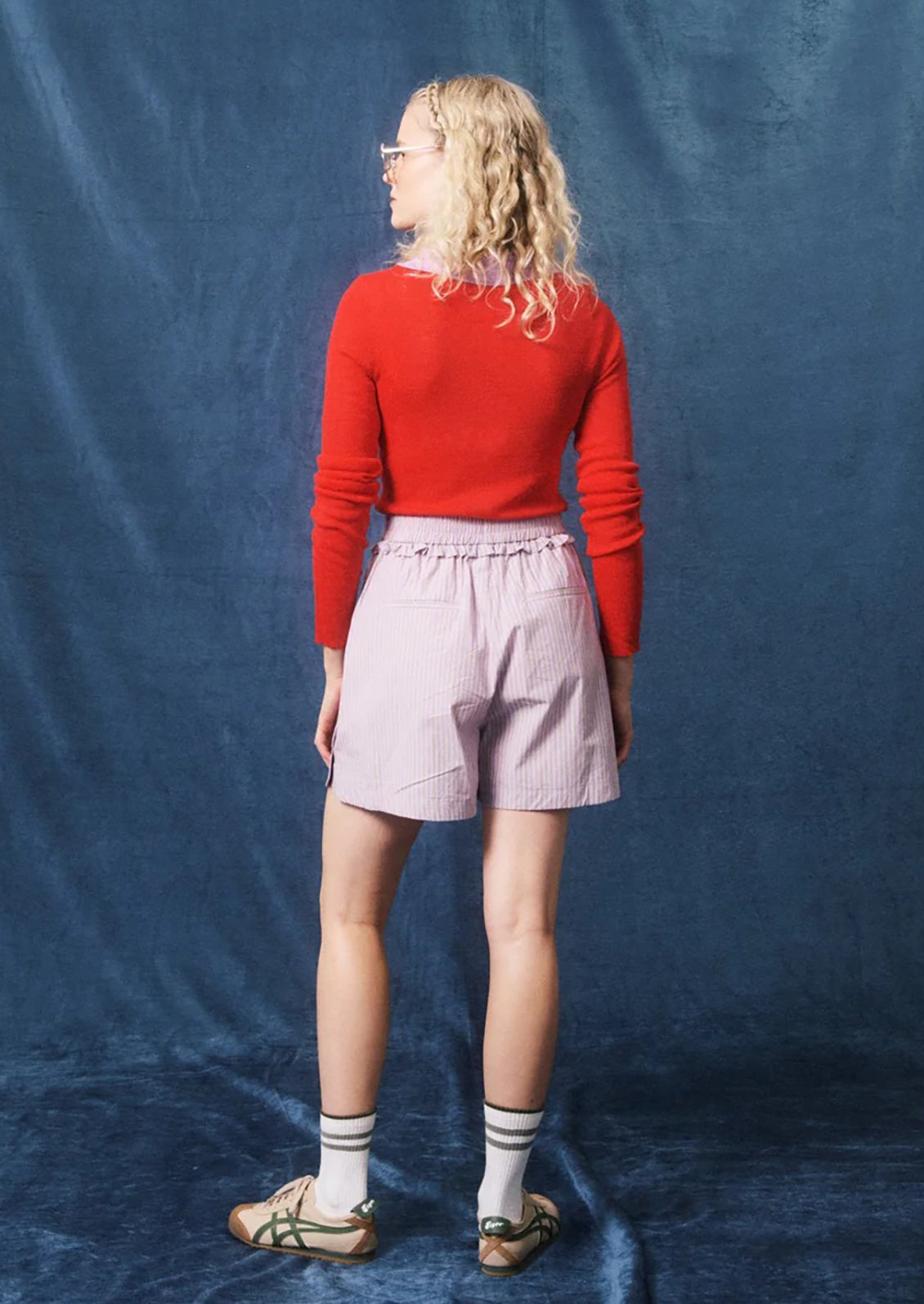 A woman stands with her back to the camera wearing a bright red long-sleeve top, light pink pleated shorts, striped socks, and sandals, posed against a textured blue fabric backdrop.