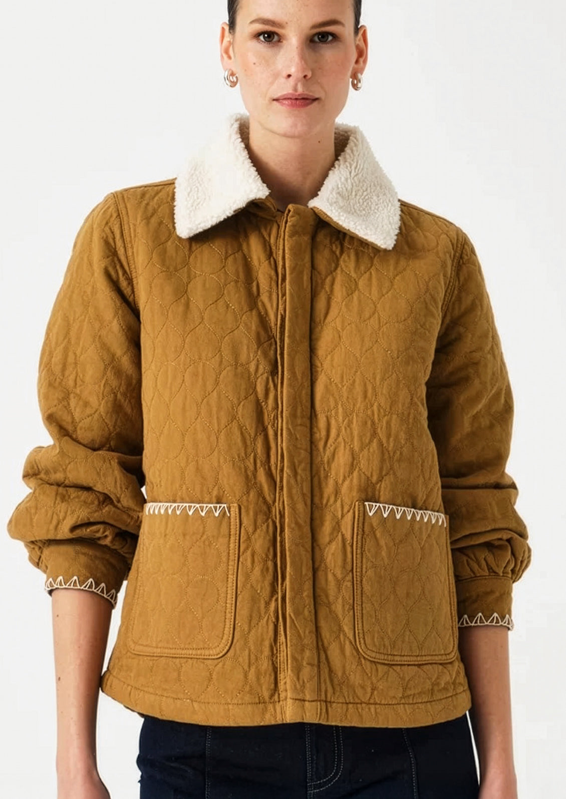 A woman wearing a quilted mustard-brown jacket with a white sherpa collar and decorative white zigzag stitching on the pockets and cuffs, photographed against a plain white background.