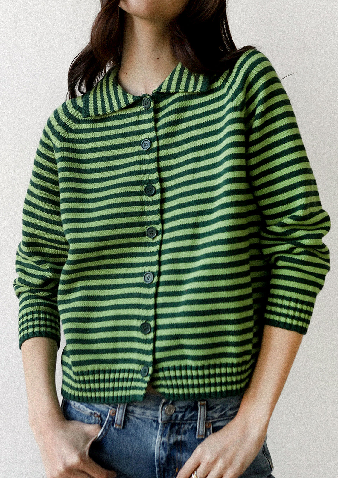 A woman wearing a green and dark green horizontally striped knit cardigan with a collar and front buttons, paired with blue jeans, against a plain light background.
