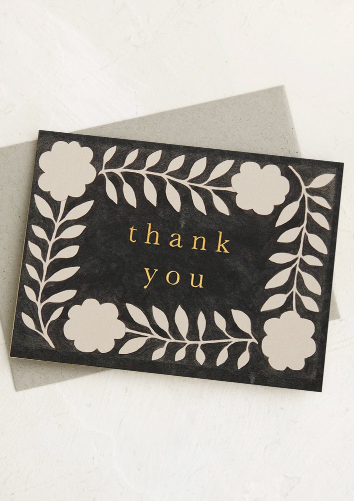 A black card with white floral pattern with "Thank you" in gold letters.